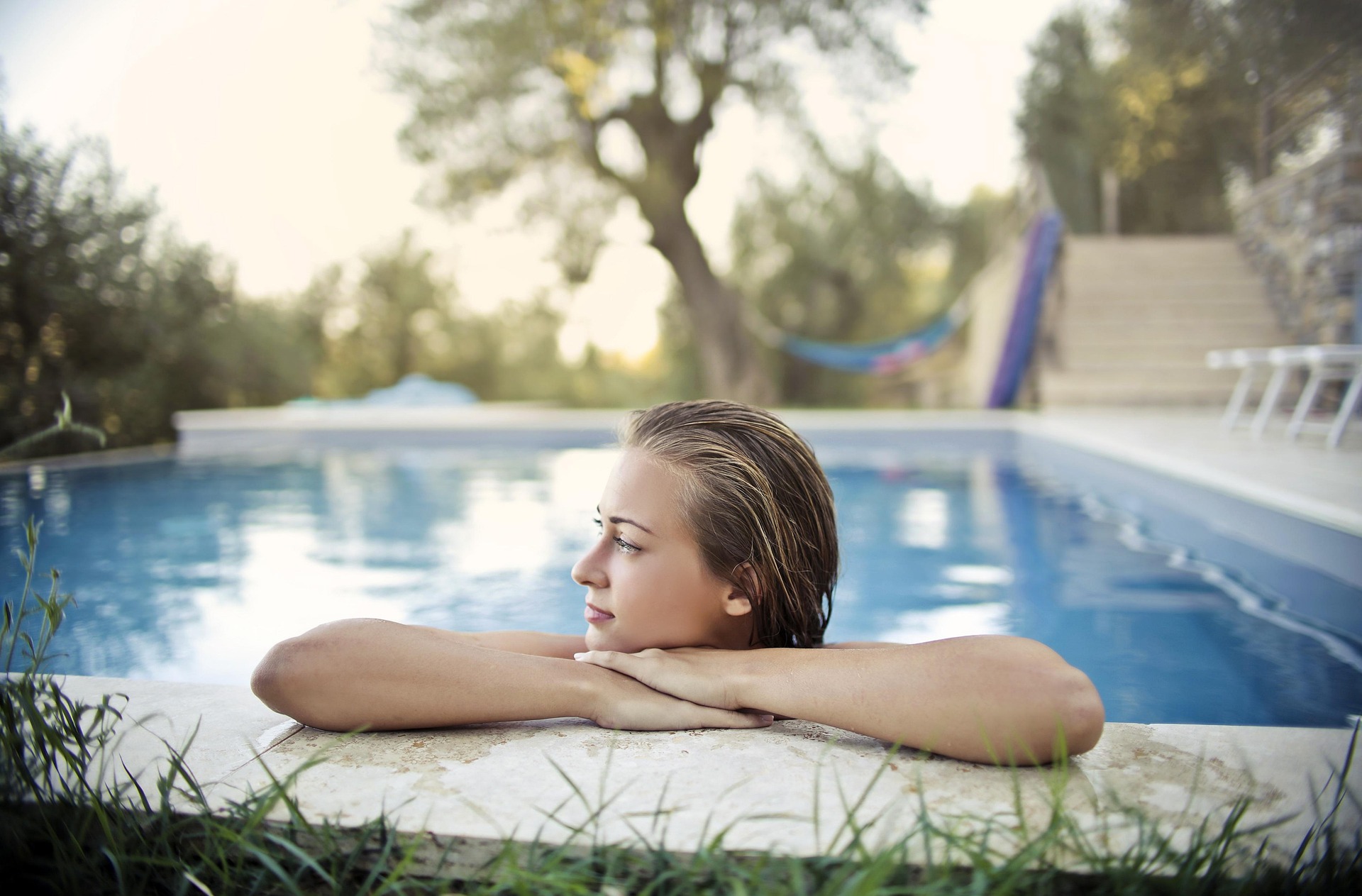 Woman relaxing in a swimming pool with her arms resting on the edge, enjoying a peaceful wellness and skin recovery moment outdoors.