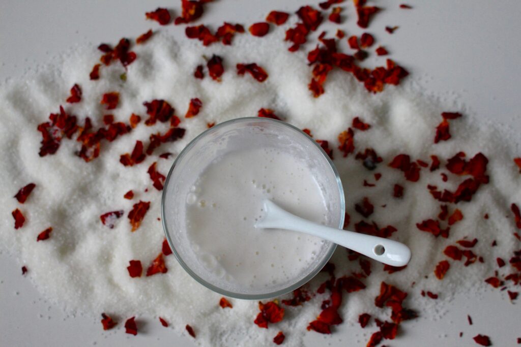 Bowl of creamy natural facial mask surrounded by bath salts and dried rose petals for a relaxing spa skincare treatment.