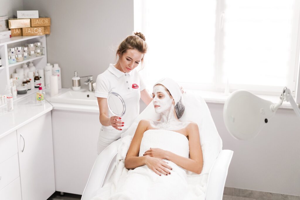 Woman receiving a professional facial mask treatment at a skincare clinic while an esthetician shows her the results in a handheld mirror.