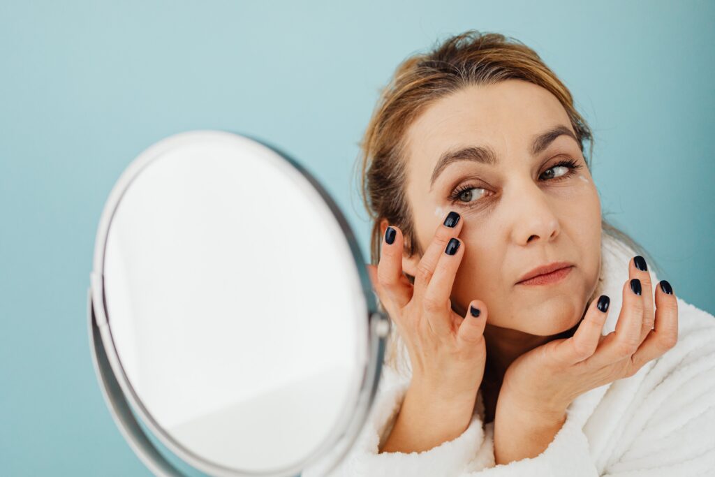 siguiente foto
Alt Text:
Woman applying under-eye cream while looking in a mirror as part of her daily skincare routine to reduce dark circles and fine lines.